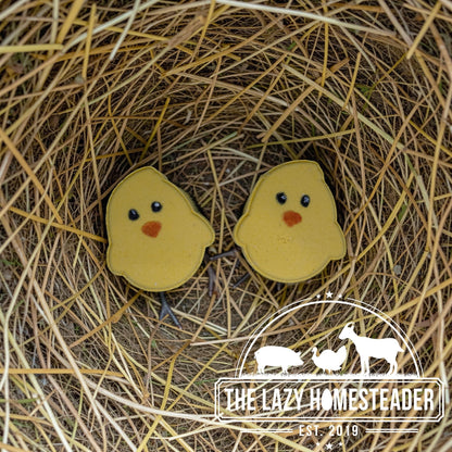 The Lazy Homesteader LLC Chick bath bomb shaped like a yellow chick with black eyes and orange beak, nestled in straw.